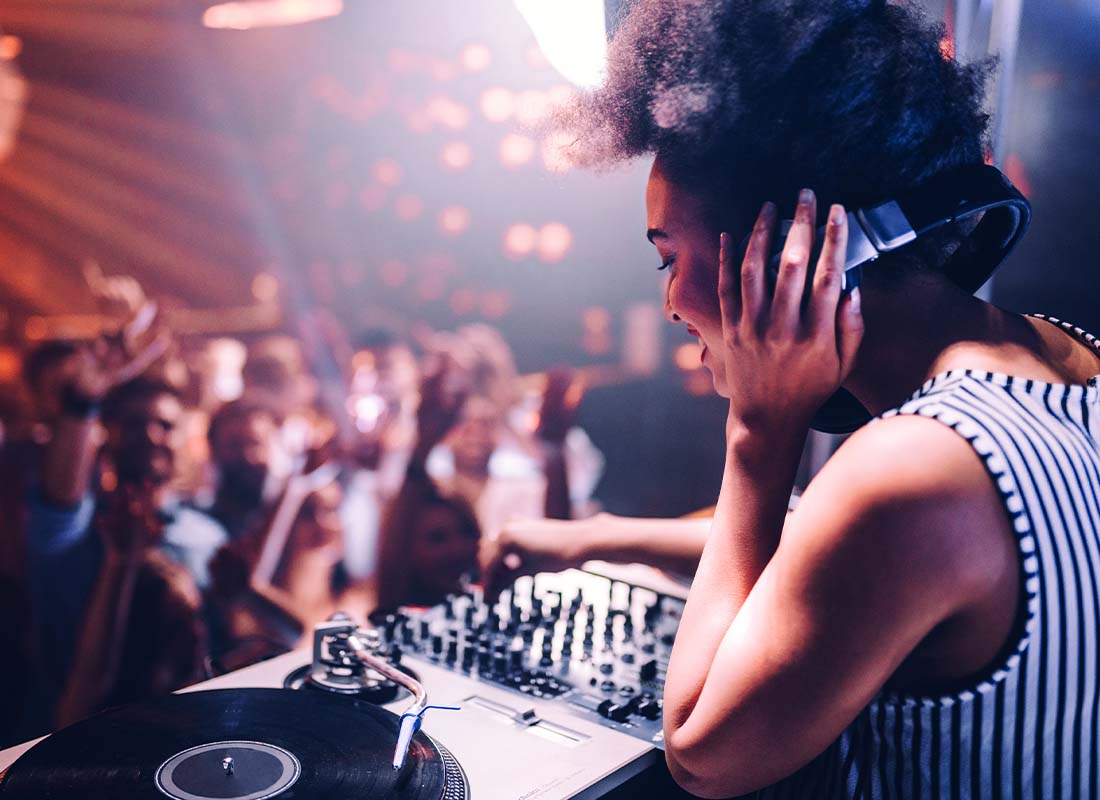 Nightclub and Bar Insurance - Focus of a DJ in a Nightclub Smiling and Listening to Her Own Beats in Front of a Large Dancing Crown with Lots of Neon Lights